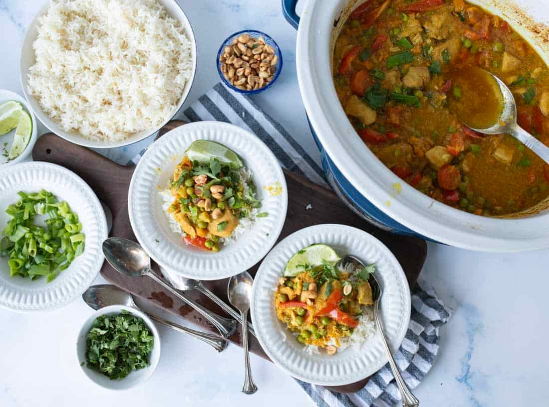 A table set with bowls of rice, a curry dish in a pot, plates of curry over rice, and small bowls of chopped green onions, cilantro, peanuts, and lime wedges, plus several serving spoons.