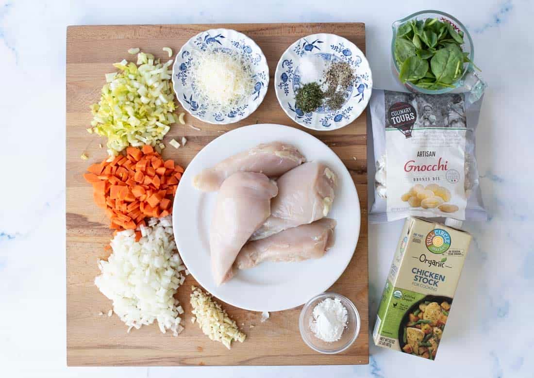 A wooden board with raw chicken breasts on a plate, chopped carrots, celery, onion, and garlic, plus small bowls of grated cheese, salt, dried herbs, and flour. Also shown are spinach leaves, packaged gnocchi, and chicken stock.
