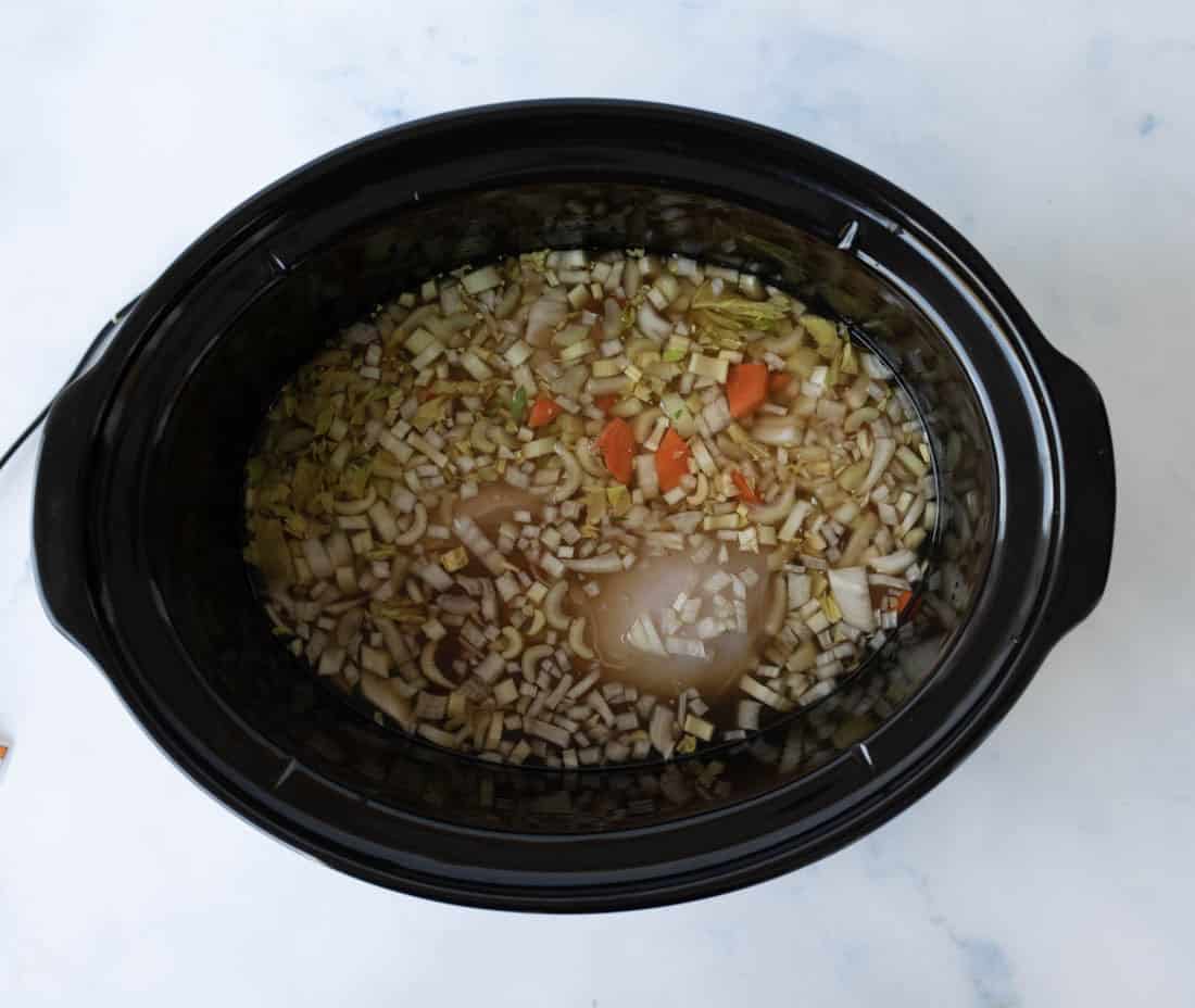 A slow cooker filled with raw chopped onions, carrots, celery, and liquid broth, with pieces of raw chicken submerged in the mixture.