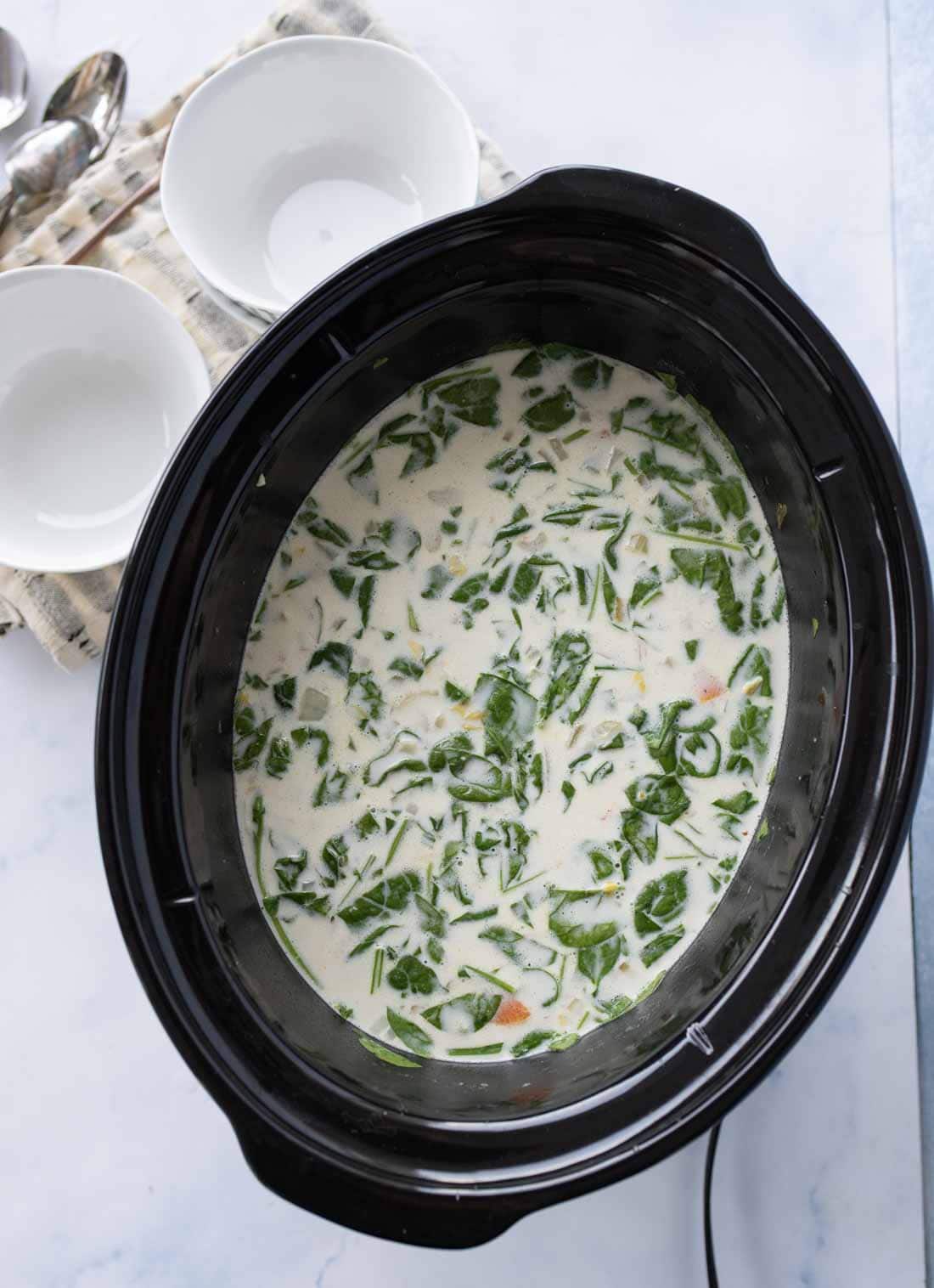 A slow cooker filled with creamy soup containing chopped spinach and vegetables, placed on a light surface next to two white bowls and a spoon on a cloth napkin.