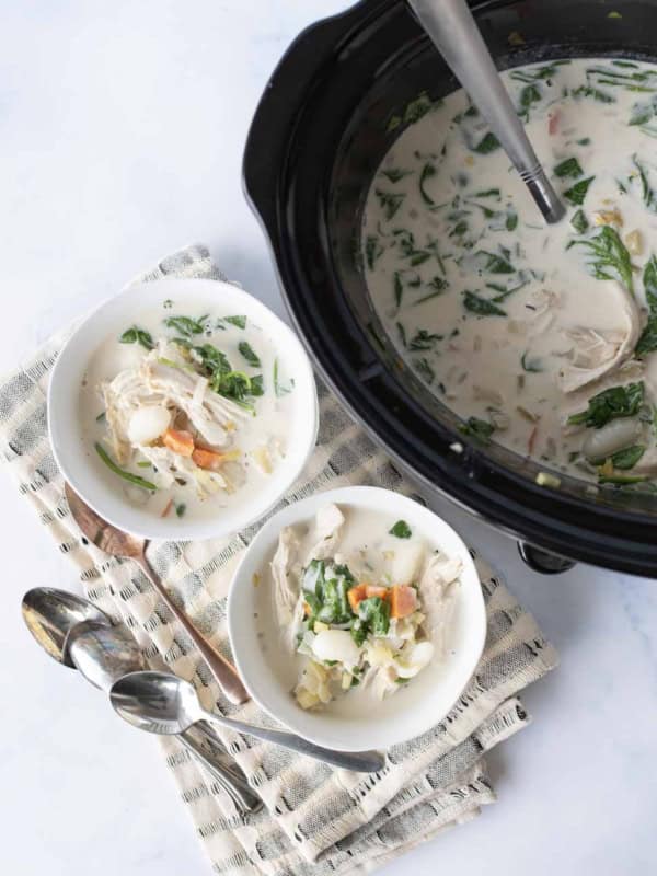 Overhead view of a black slow cooker filled with creamy soup, next to two white bowls of soup with chicken, vegetables, and spinach, all placed on a light cloth with two metal spoons beside them.
