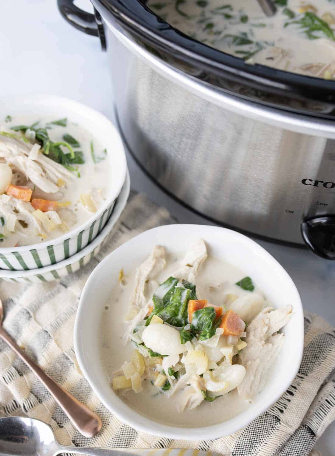 Two bowls of creamy italian chicken gnocchi soup sit on a cloth next to a slow cooker filled with more soup. Visible ingredients include shredded chicken, carrots, white beans, spinach, and a creamy broth.