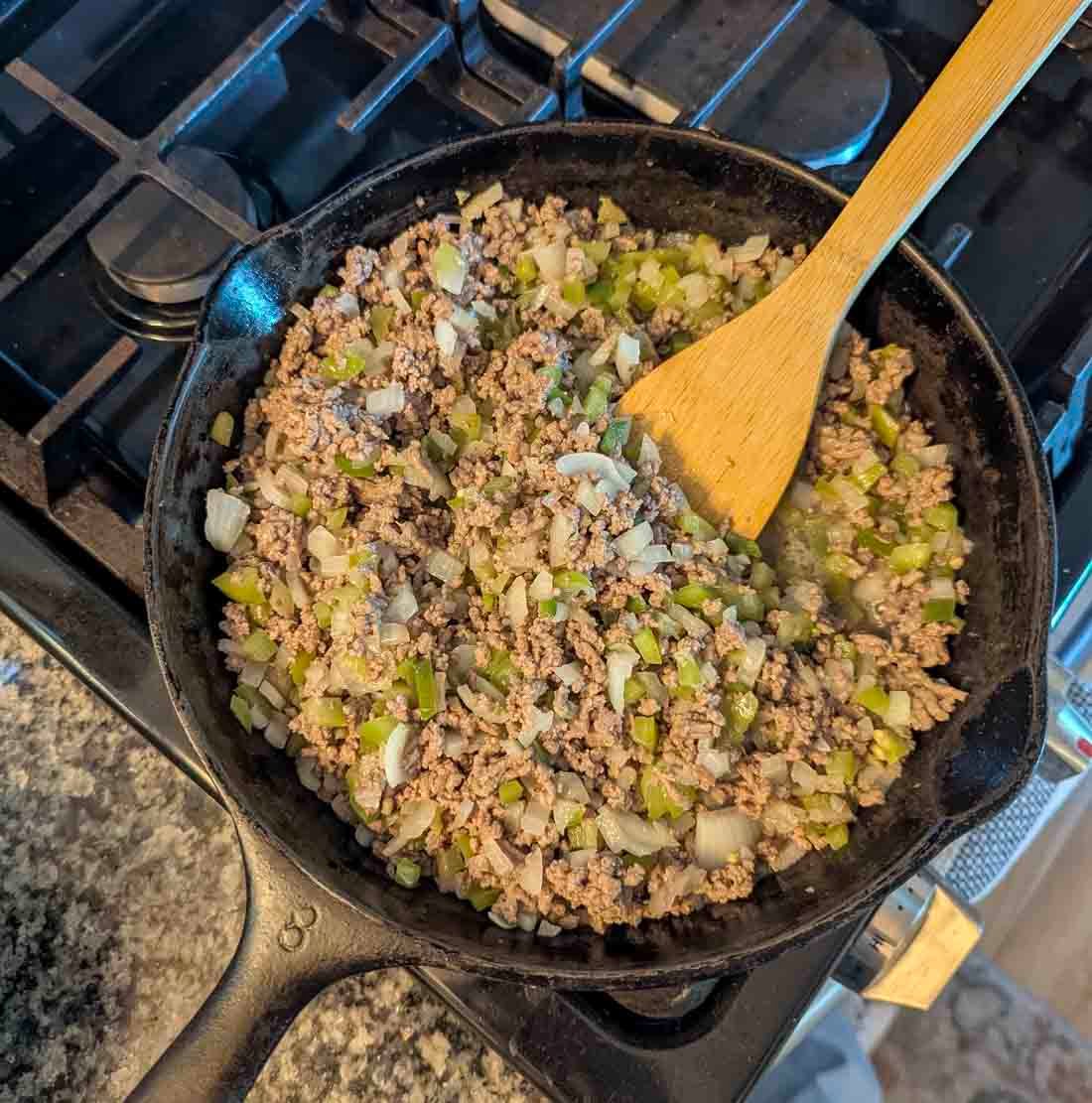 A cast iron skillet on a stove contains cooked ground beef mixed with chopped onions and green peppers. A wooden spoon is resting in the skillet, stirring the mixture.
