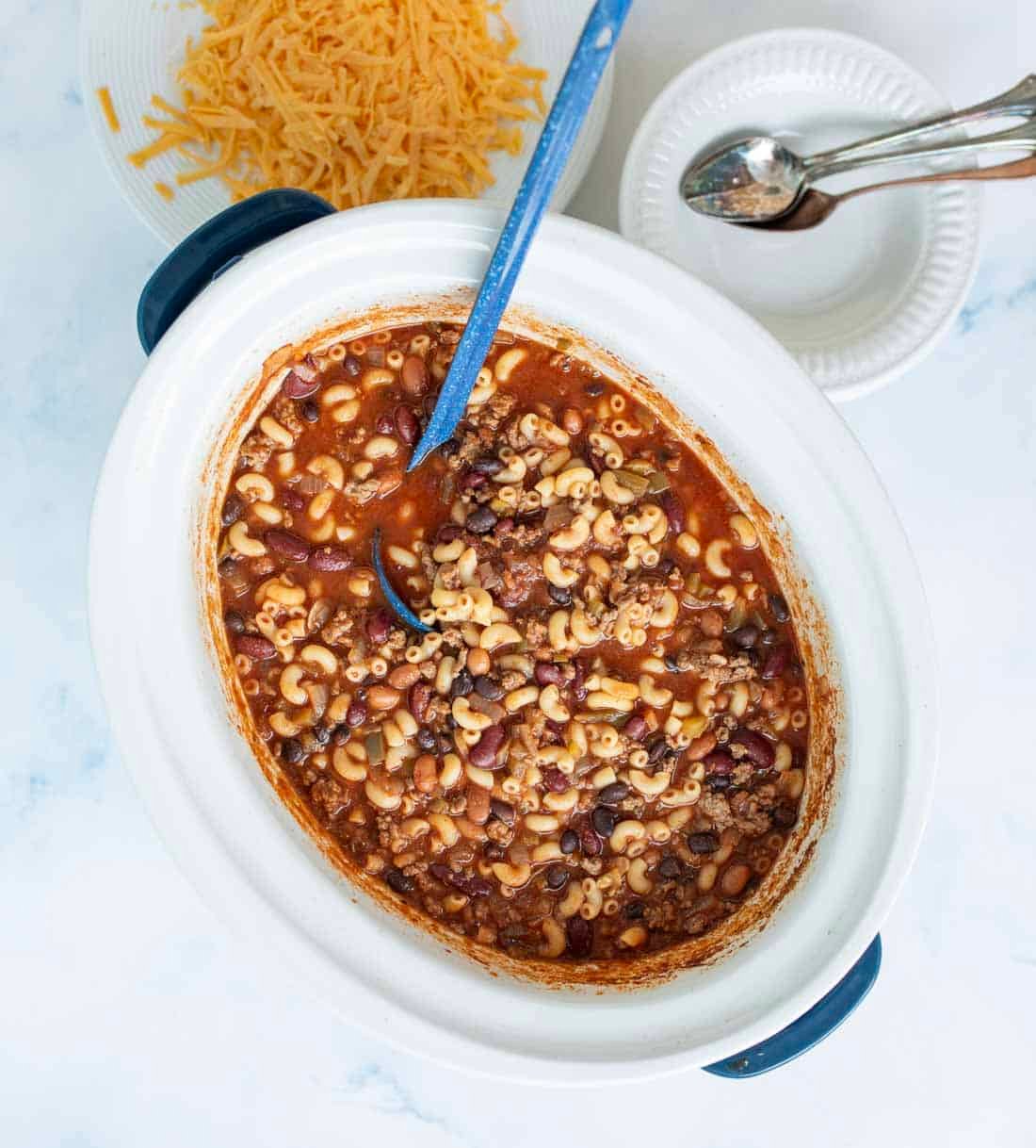 A slow cooker filled with chili macaroni, featuring ground beef, beans, macaroni noodles, and tomato sauce. A blue spoon rests inside, with shredded cheese and stacked white plates with spoons nearby.