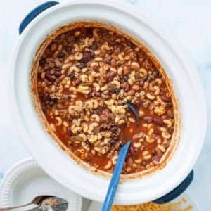A slow cooker filled with chili mac, showing macaroni, ground beef, beans, and tomato sauce, with a blue spoon inside. Plates, shredded cheese, and spoons are nearby on a white surface.