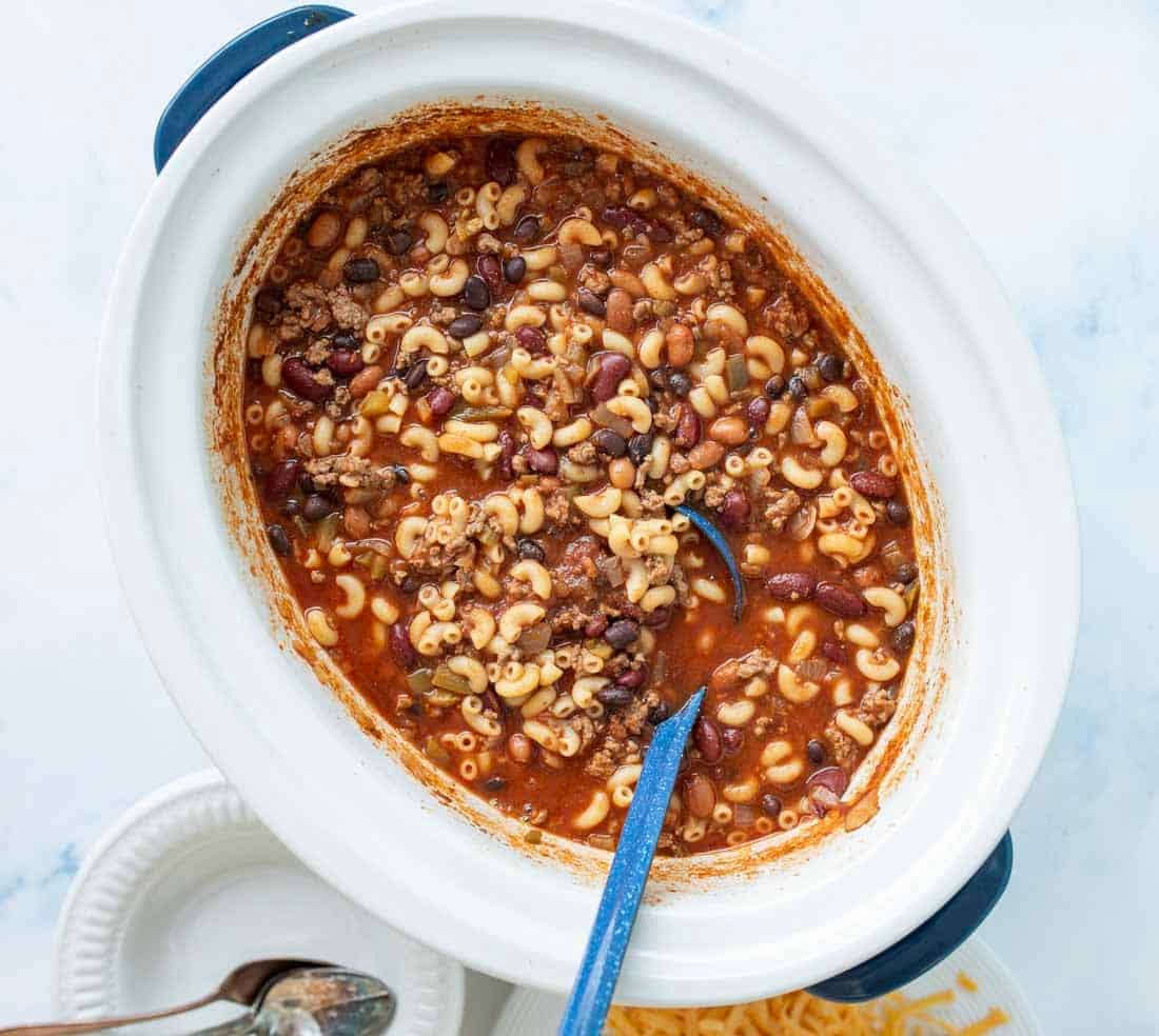 A slow cooker filled with chili mac, showing macaroni, ground beef, beans, and tomato sauce, with a blue spoon inside. Plates, shredded cheese, and spoons are nearby on a white surface.