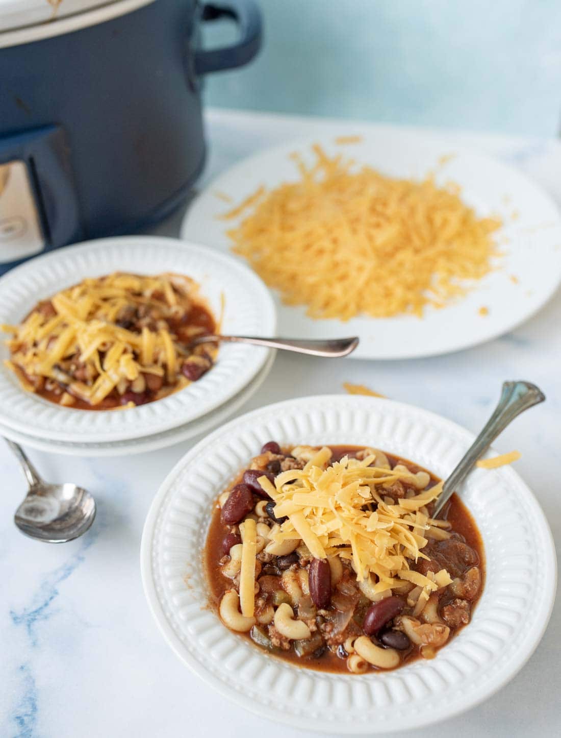 Two white bowls of slow cooker chili mac  with chili, macaroni, and kidney beans, topped with shredded cheddar cheese, on a white surface. A plate of extra grated cheese and a slow cooker are in the background.