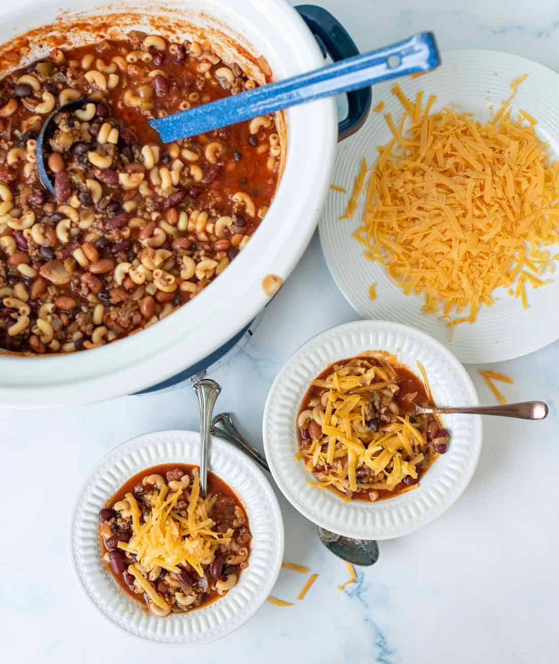 A crockpot filled with chili containing macaroni, beans, and ground meat sits beside a plate of shredded cheddar cheese and two bowls of chili topped with cheese on a white surface. Spoons are placed in the bowls.