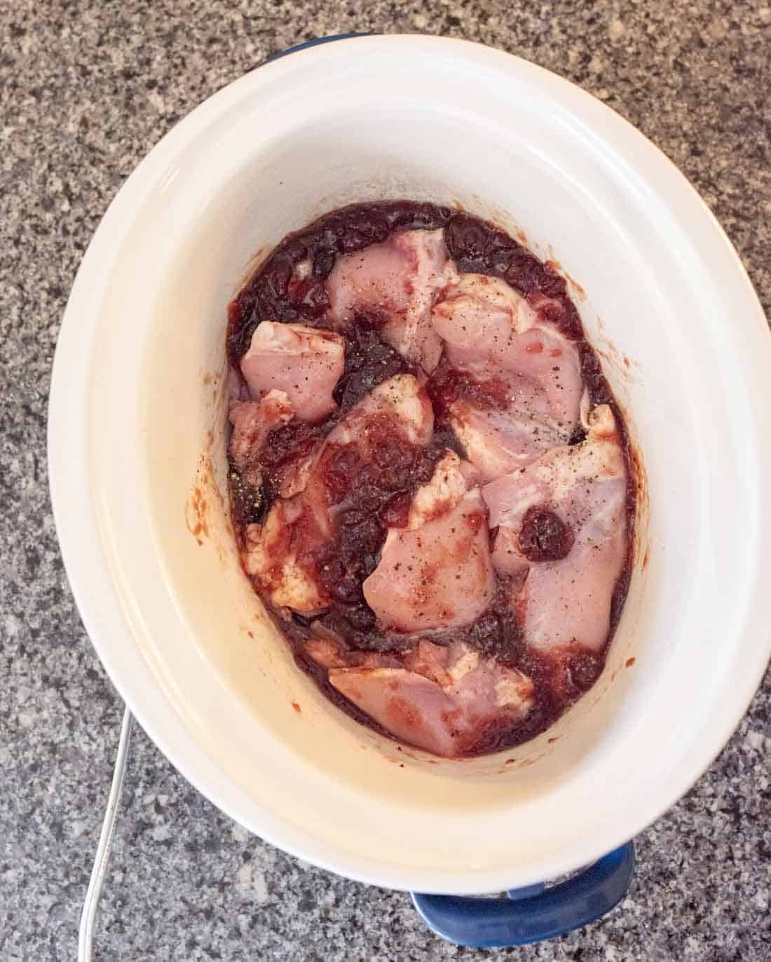 Raw chicken pieces with a dark red sauce sit in a white slow cooker on a gray speckled countertop, ready to be cooked.
