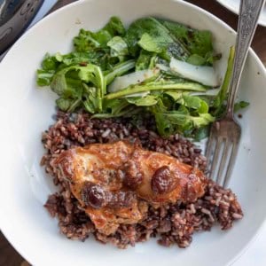 A white plate with cranberry chicken topped with sauce and cherries on a bed of rice, served alongside a green leafy salad with cucumber slices. A fork rests on the plate.