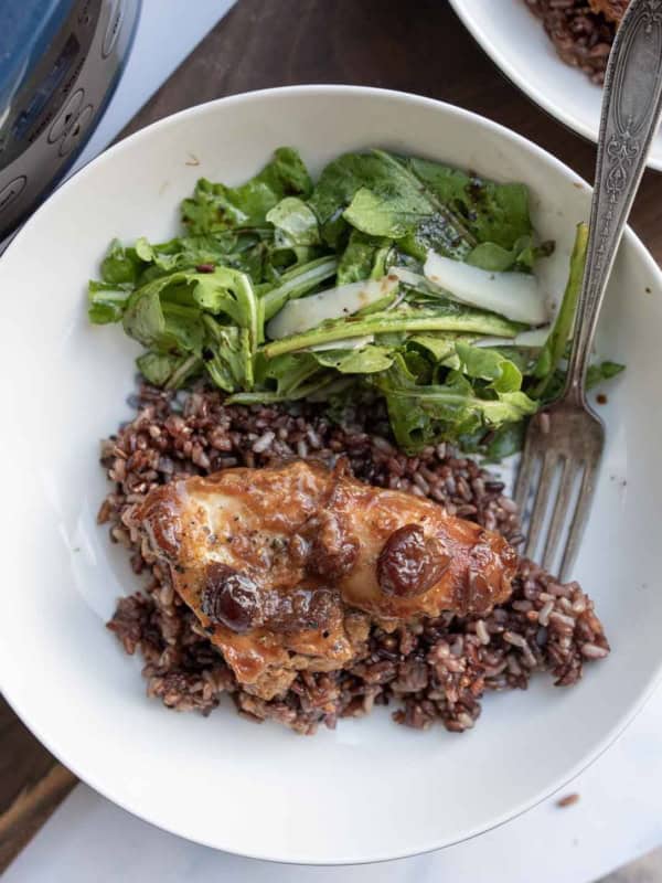 A white plate with cranberry chicken topped with sauce and cherries on a bed of rice, served alongside a green leafy salad with cucumber slices. A fork rests on the plate.