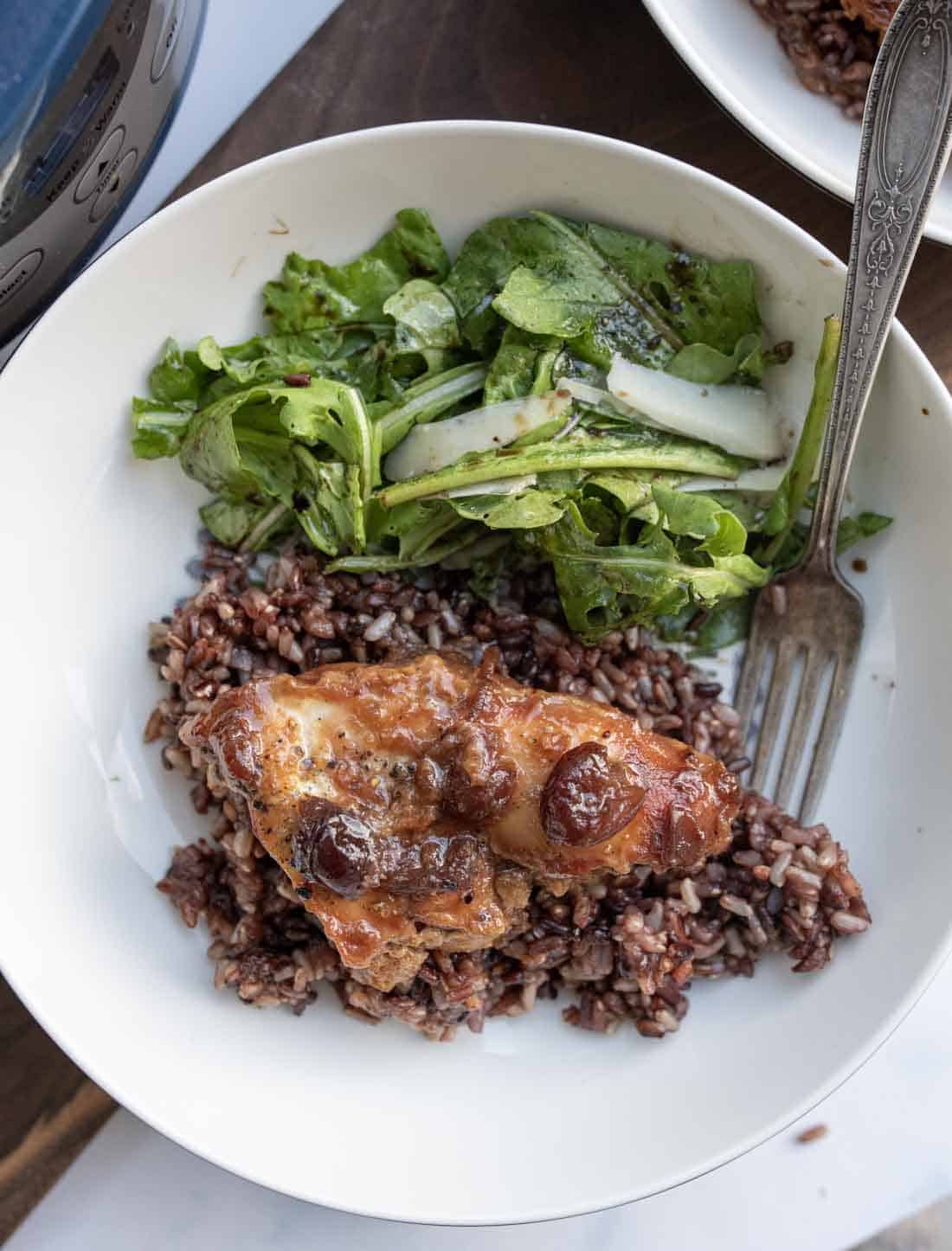 A white plate with cranberry chicken topped with sauce and cherries on a bed of rice, served alongside a green leafy salad with cucumber slices. A fork rests on the plate.