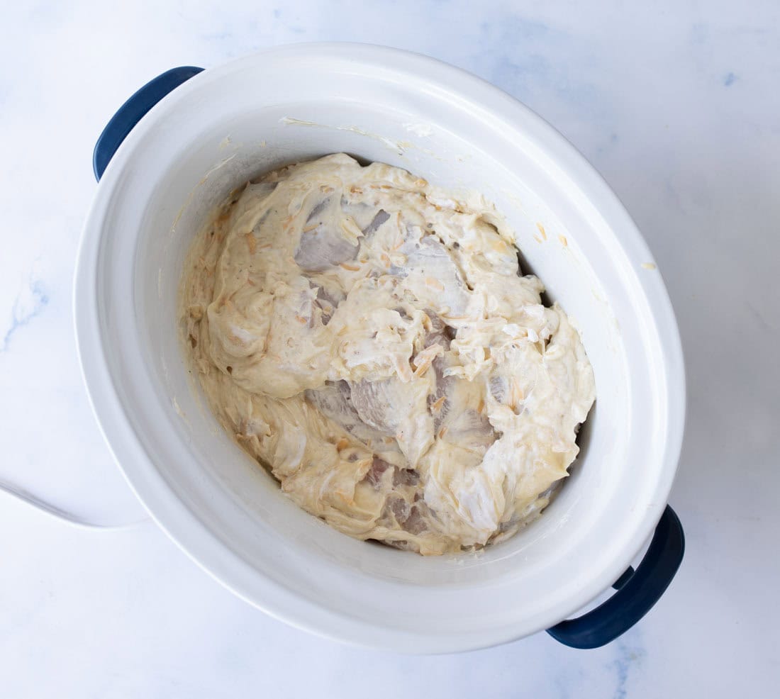 Raw chicken breasts covered in a creamy, seasoned mixture inside a white slow cooker with dark blue handles, placed on a light marble surface.