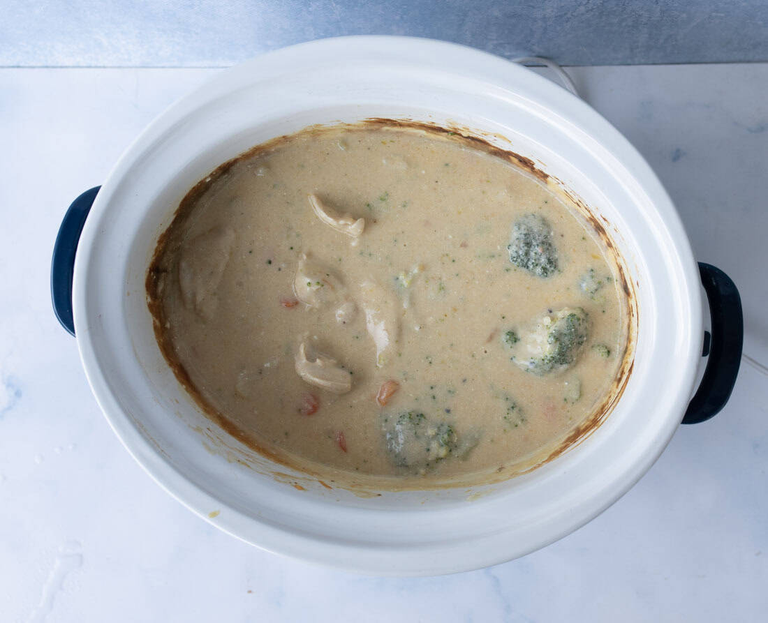 A slow cooker filled with creamy soup containing pieces of chicken, broccoli florets, and sliced carrots, sitting on a light-colored surface.