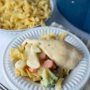 A white plate with cooked egg noodles topped with slow cooker creamy chicken, sliced carrots, broccoli, and cauliflower. In the background, a bowl of plain egg noodles and part of a blue pot are visible.