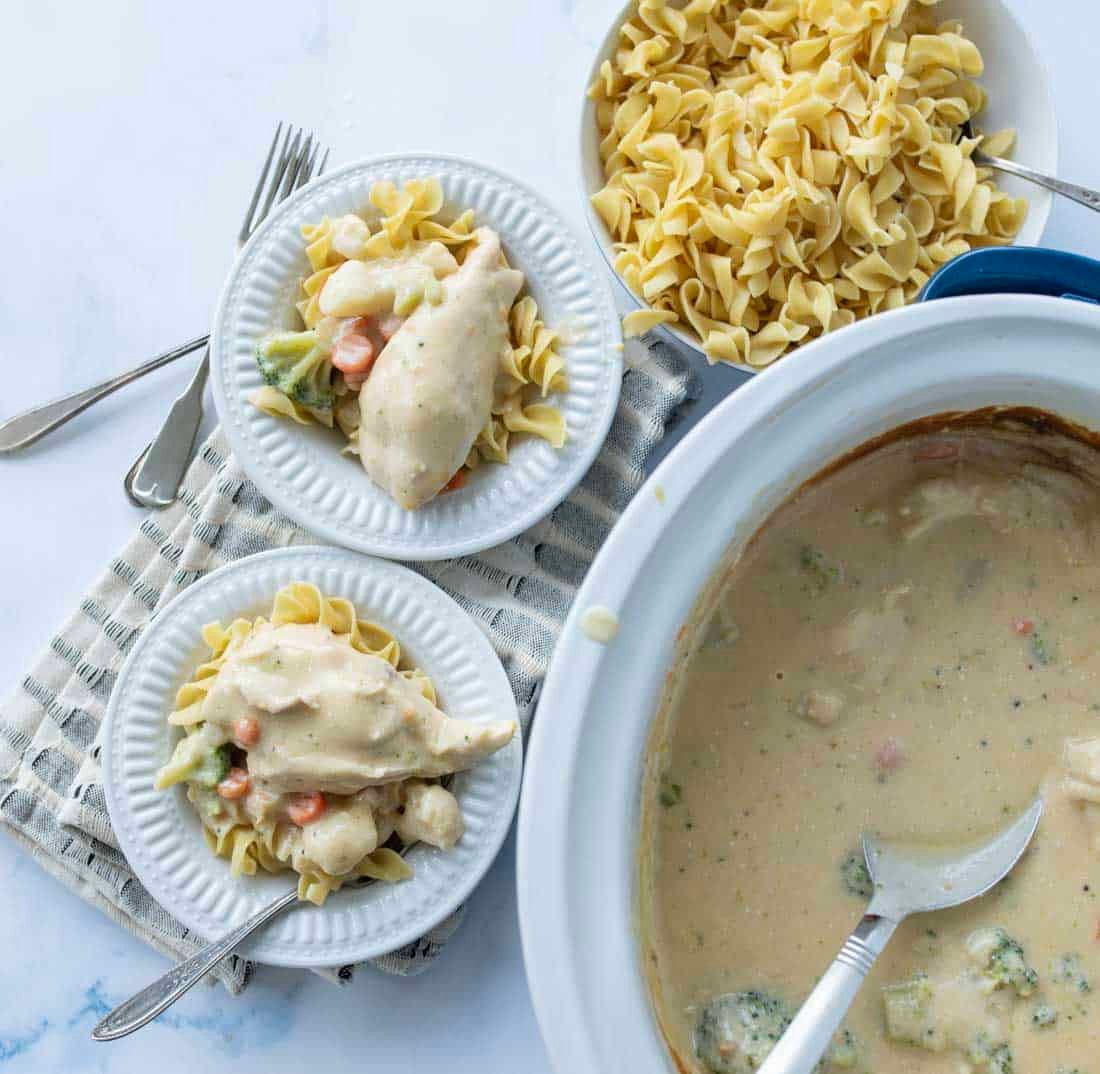 Two plates of chicken and vegetables in creamy sauce served over egg noodles, next to a large bowl of plain noodles and a slow cooker with remaining creamy chicken mixture. A fork and two knives lie nearby on a striped cloth.