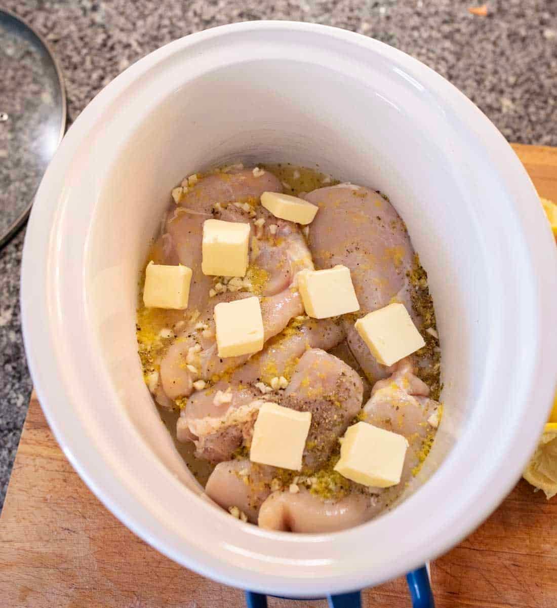 Raw chicken thighs seasoned with herbs and minced garlic, topped with cubes of butter, placed in a white slow cooker on a wooden board with a granite countertop in the background.