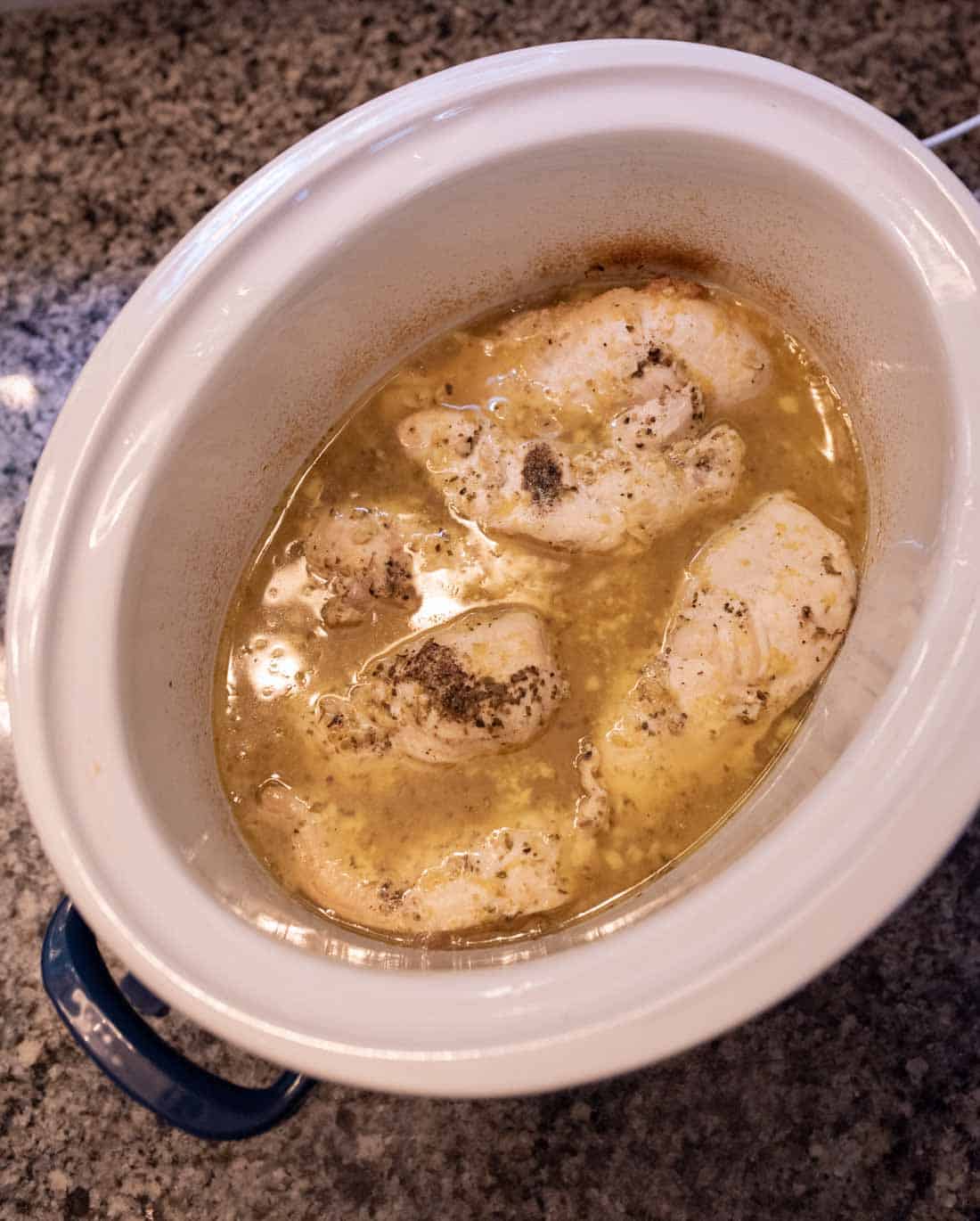 Chicken breasts simmer in a seasoned broth inside a white slow cooker, set on a speckled granite countertop.