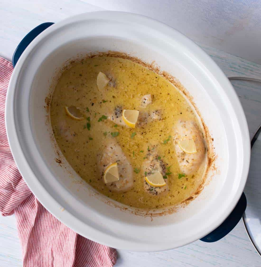 A slow cooker filled with creamy lemon chicken, garnished with lemon wedges and chopped herbs. The cooker sits on a white surface beside a pink-striped cloth.