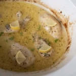 Four chicken breasts sit in a creamy, seasoned sauce inside a white slow cooker, each piece topped with a lemon wedge and a sprinkle of herbs. The sauce has a slightly browned edge around the cooker.