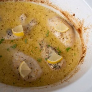Four chicken breasts sit in a creamy, seasoned sauce inside a white slow cooker, each piece topped with a lemon wedge and a sprinkle of herbs. The sauce has a slightly browned edge around the cooker.