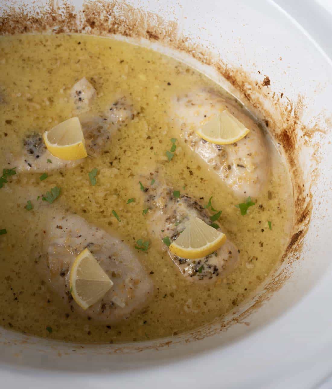 Four chicken breasts sit in a creamy, seasoned sauce inside a white slow cooker, each piece topped with a lemon wedge and a sprinkle of herbs. The sauce has a slightly browned edge around the cooker.