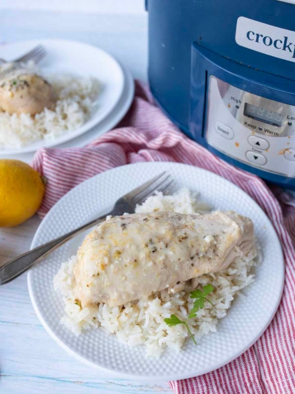 A plate of white rice topped with slow cooker lemon chicken, garnished with herbs, sits next to a fork and knife. A slow cooker and lemon are in the background on a red striped napkin.