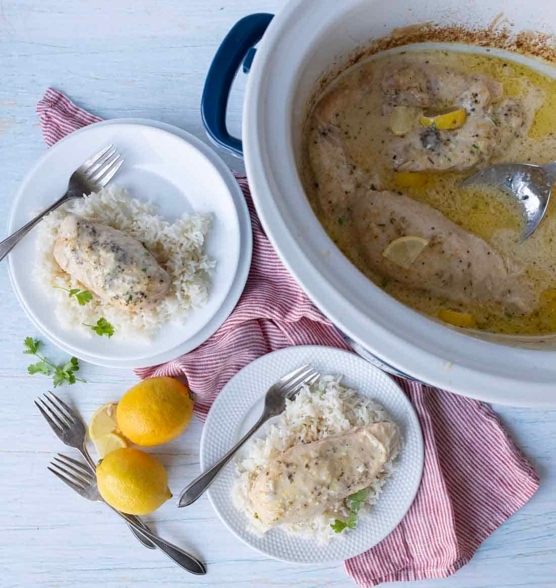 Overhead view of creamy lemon chicken with sauce in a slow cooker, served on plates with white rice. Two plates are on a red-striped cloth, with fresh lemons and forks nearby.
