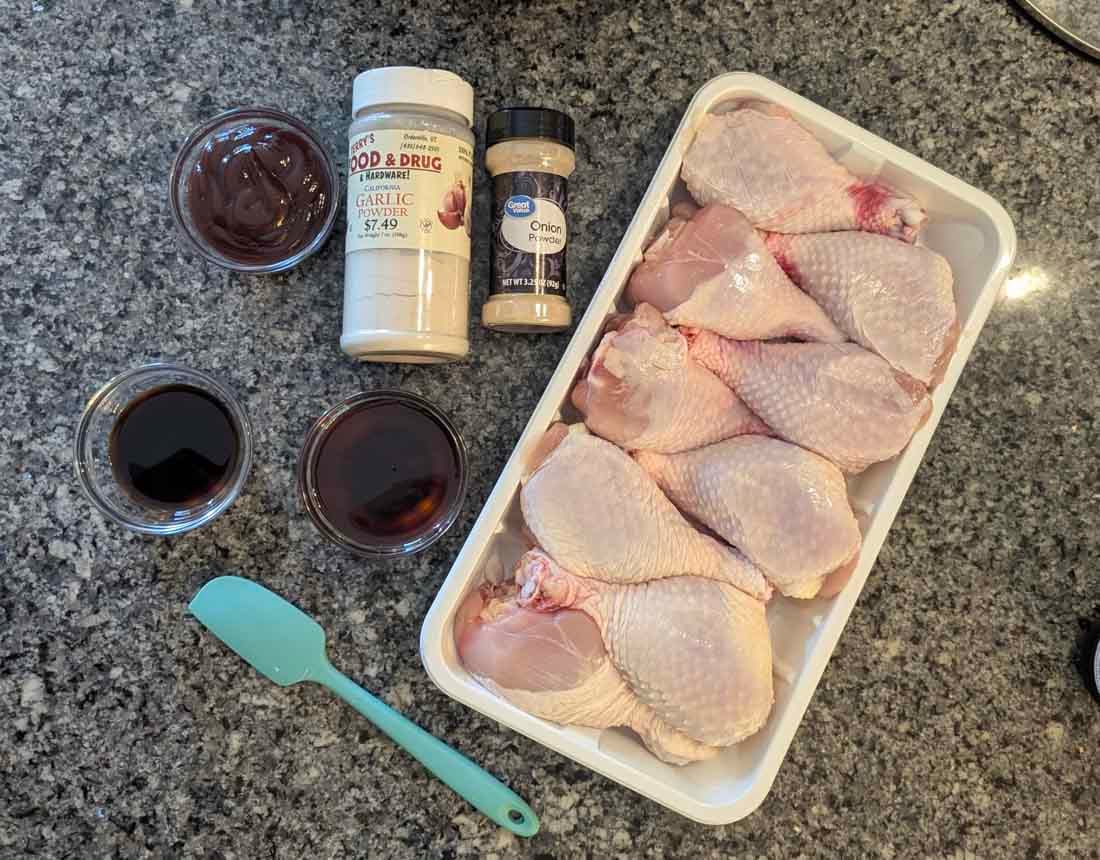 Raw chicken drumsticks in a plastic tray, with small bowls of barbecue sauce and soy sauce, a teal spatula, and containers of garlic powder and onion powder on a granite countertop.