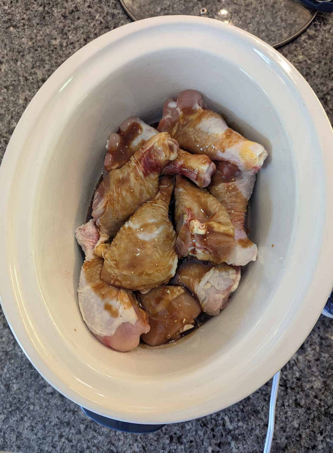 Raw chicken drumsticks coated in a marinade are placed inside a white slow cooker, which sits on a gray granite countertop.