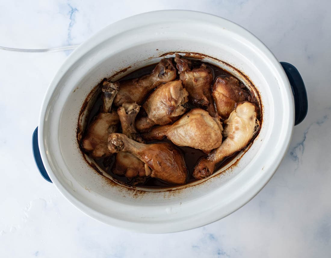 A white slow cooker filled with cooked chicken drumsticks in a dark sauce, viewed from above on a light marble surface.