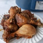 A white plate filled with cooked slow cooker chicken legs sits on a light surface, with a blue slow cooker visible in the background.