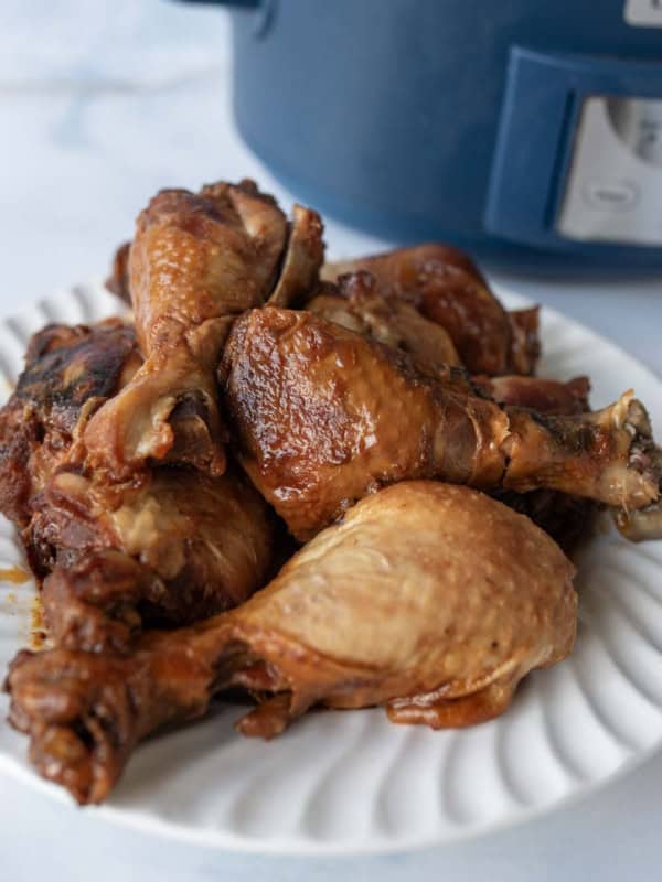 A white plate filled with cooked slow cooker chicken legs sits on a light surface, with a blue slow cooker visible in the background.