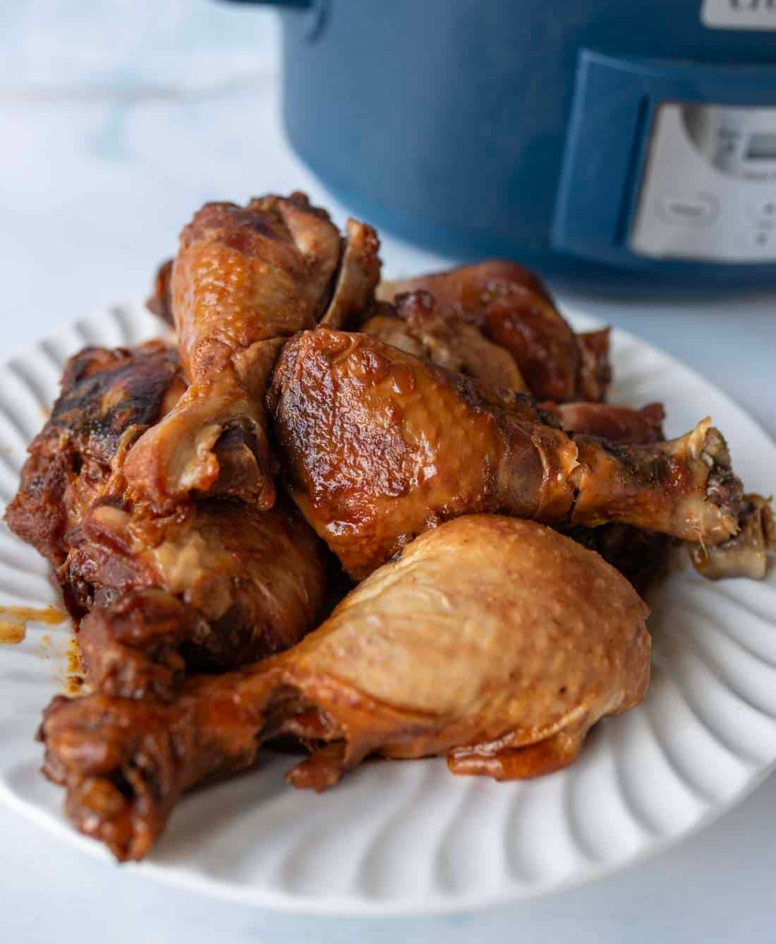 A white plate filled with cooked slow cooker chicken legs sits on a light surface, with a blue slow cooker visible in the background.