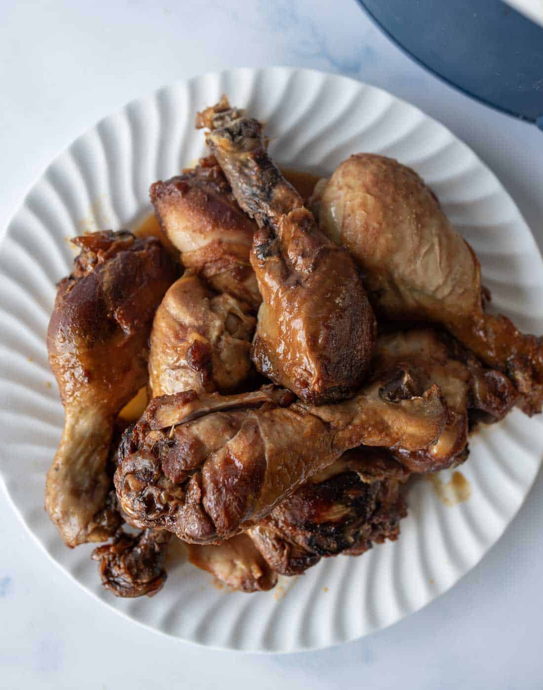 A white plate filled with several cooked chicken drumsticks, arranged in a pile on a light-colored surface.