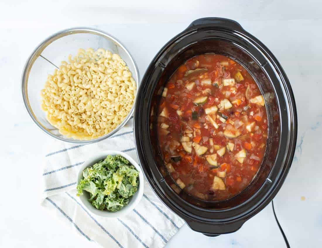 A slow cooker filled with a tomato-based soup with chopped vegetables sits next to a bowl of cooked macaroni and a small bowl of leafy greens on a striped cloth.