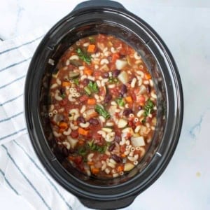 A slow cooker filled with minestrone soup featuring macaroni noodles, kidney beans, diced tomatoes, carrots, potatoes, kale, and broth, sitting on a white surface next to a striped towel.