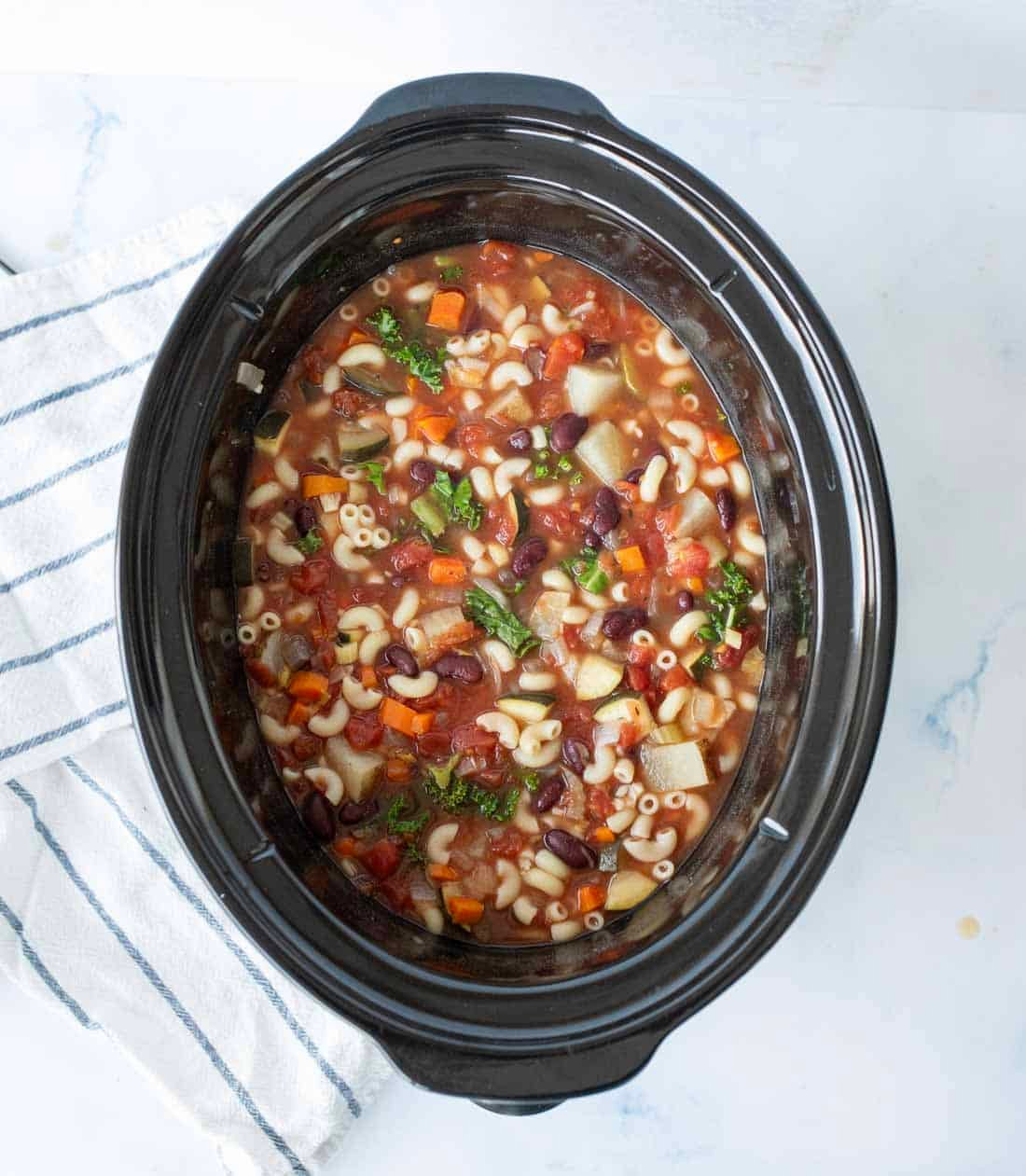 A slow cooker filled with minestrone soup featuring macaroni noodles, kidney beans, diced tomatoes, carrots, potatoes, kale, and broth, sitting on a white surface next to a striped towel.