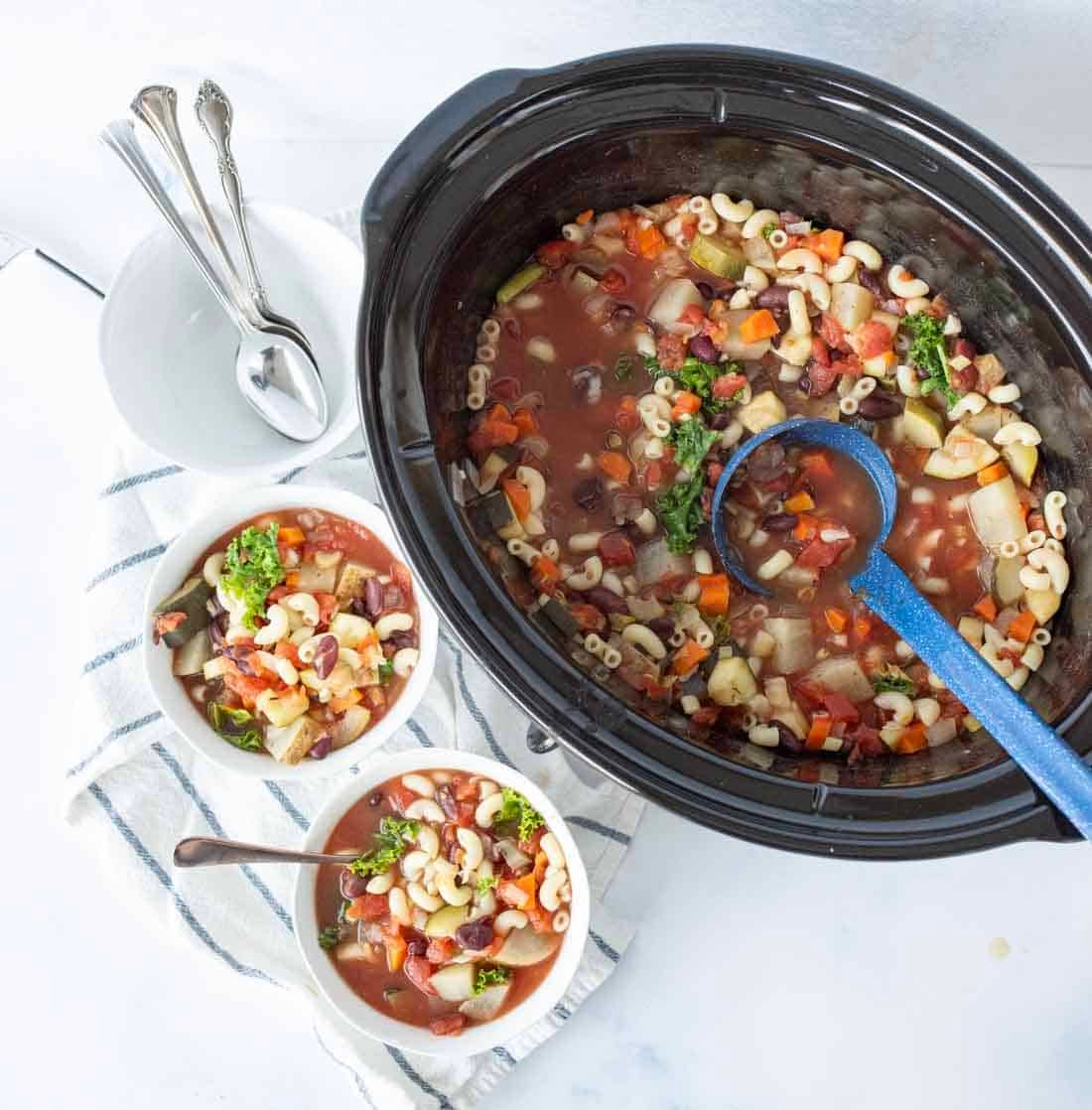 A slow cooker filled with vegetable macaroni soup, a blue ladle inside. Two bowls of the soup are served nearby on a striped towel, with spoons and an empty bowl beside them.