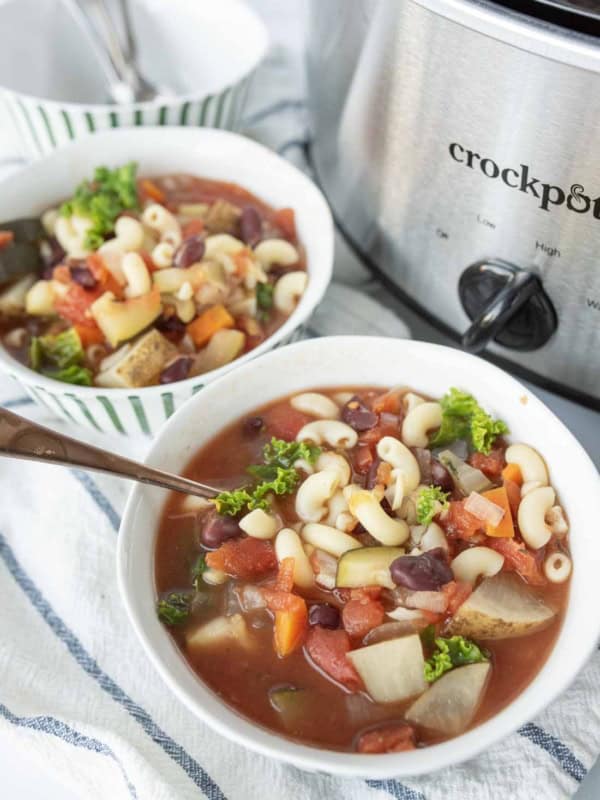 Two bowls of slow cooker minestrone soup with pasta, beans, carrots, potatoes, and kale sit on a striped towel next to a stainless steel Crockpot. One bowl has a spoon resting in it.