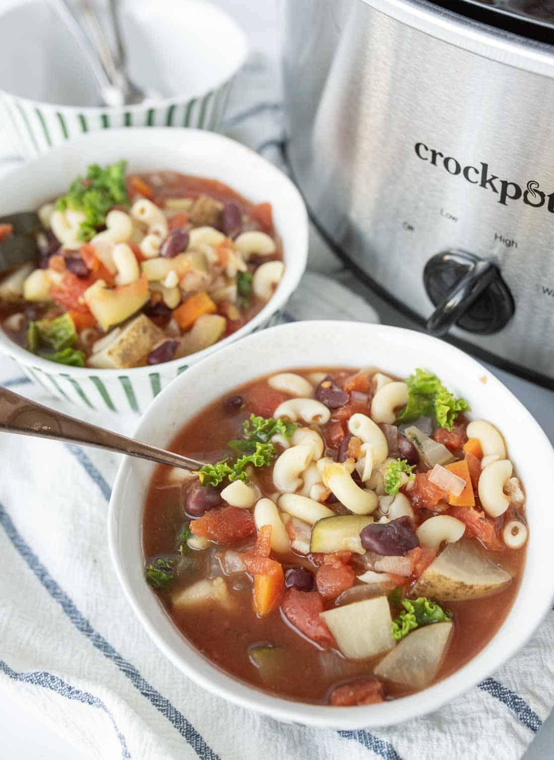 Two bowls of slow cooker minestrone soup with pasta, beans, carrots, potatoes, and kale sit on a striped towel next to a stainless steel Crockpot. One bowl has a spoon resting in it.