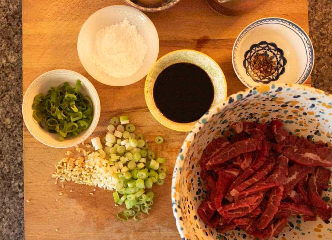 A wooden board with sliced green onions, chopped garlic, a bowl of sliced raw beef, soy sauce, cornstarch, and a small bowl of chopped dried chili peppers arranged for cooking.