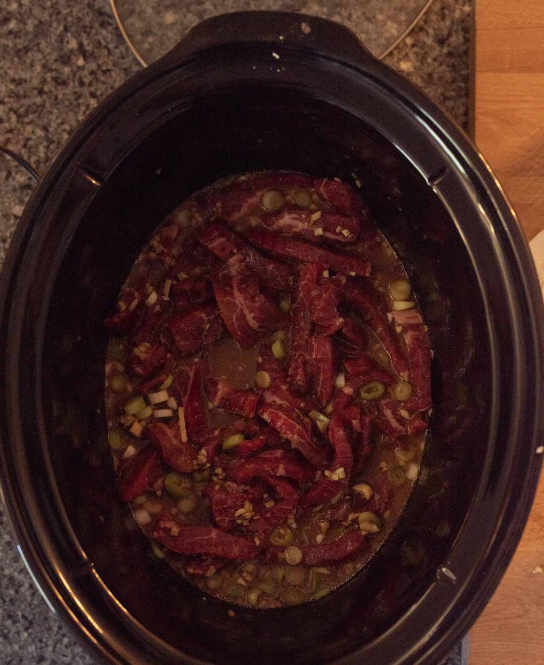 Strips of raw beef, chopped green onions, and seasonings in broth inside a black slow cooker, placed on a countertop.