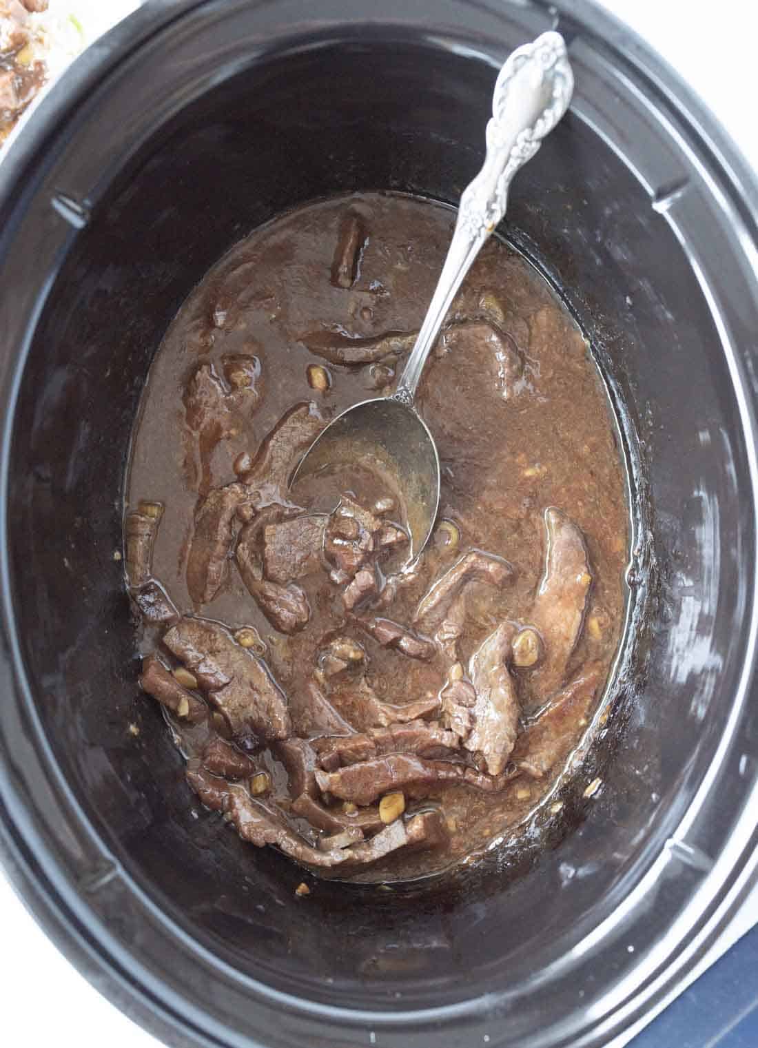 Sliced beef and mushrooms in a rich brown sauce inside a black slow cooker, with a silver spoon resting in the mixture.