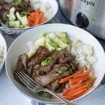 A bowl of white rice topped with sliced beef, julienned carrots, cucumber salad, and green onions sits next to a Crockpot, with a fork resting in the bowl.