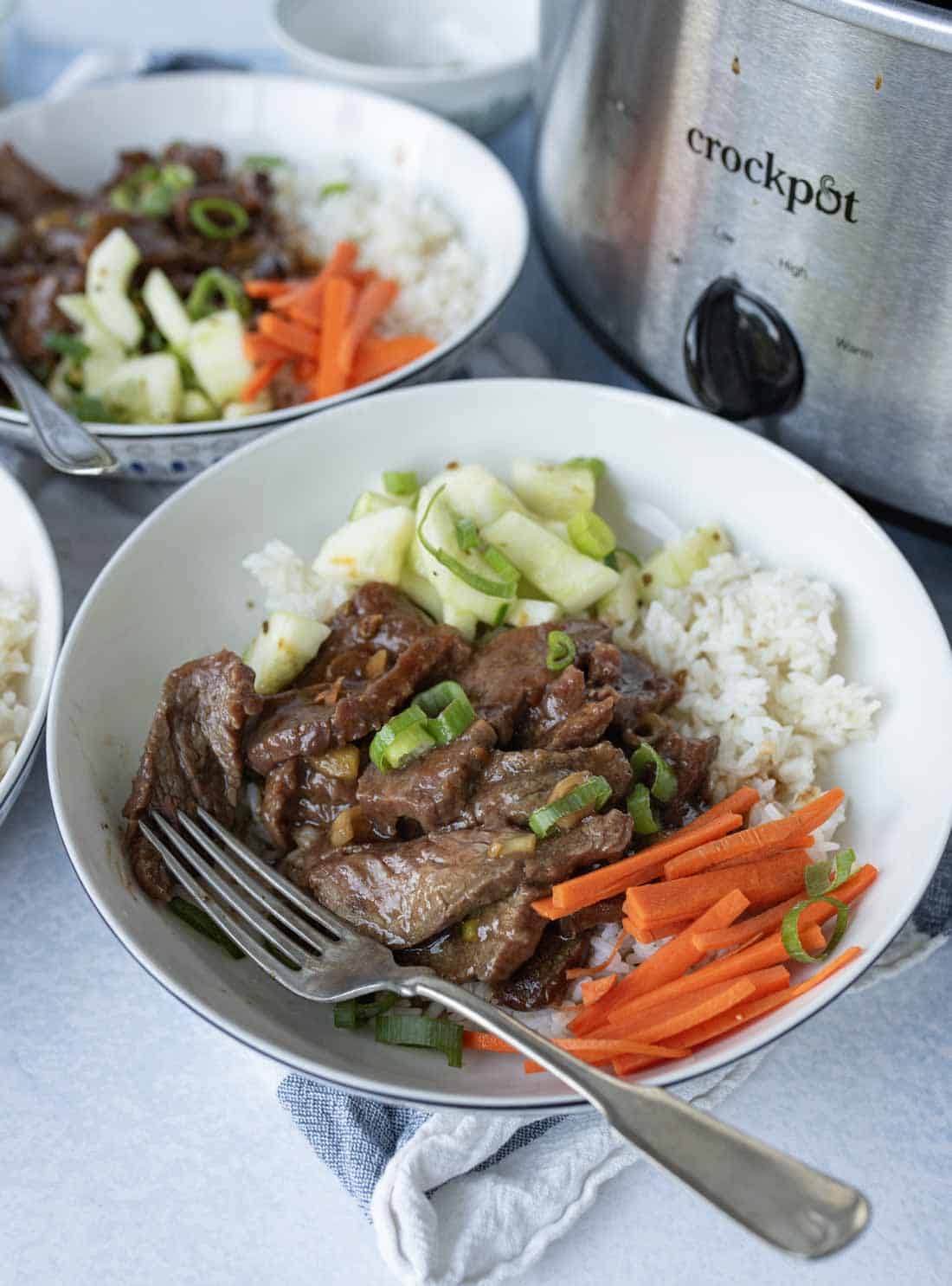 A bowl of white rice topped with sliced beef, julienned carrots, cucumber salad, and green onions sits next to a Crockpot, with a fork resting in the bowl.