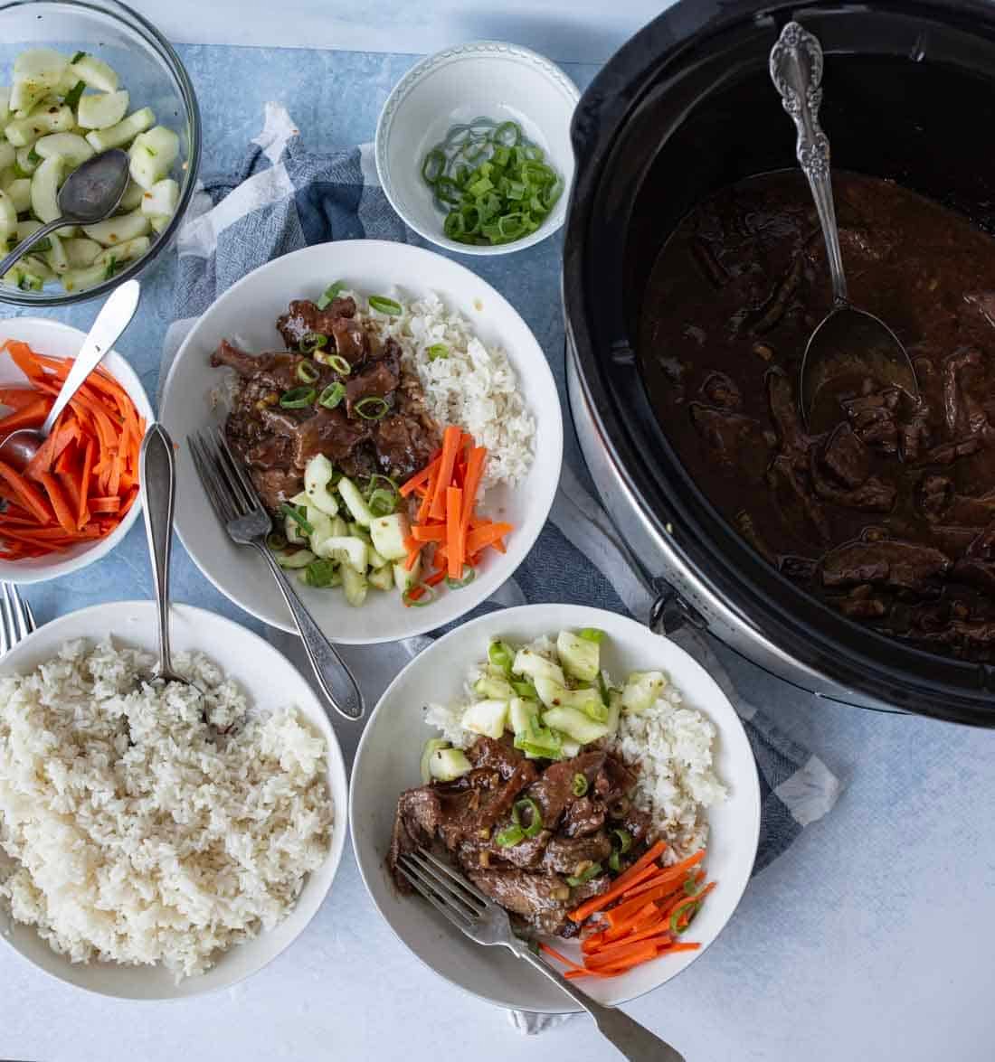 Two bowls of rice topped with beef, sliced cucumbers, carrots, and green onions sit next to a bowl of extra rice, a bowl of sliced cucumbers, and a slow cooker filled with beef in sauce. Spoons and forks are placed nearby.
