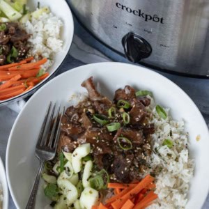 A bowl of rice topped with sliced slow cooker mongolian beef, shredded carrots, cucumbers, and green onions sits next to a Crockpot filled with more beef. A fork rests in the bowl and sesame seeds are sprinkled on top.