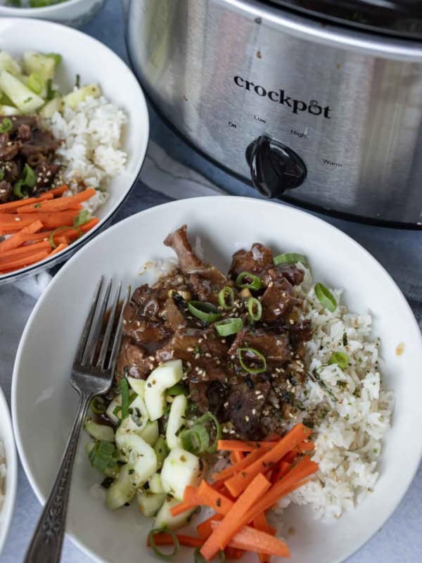 A bowl of rice topped with sliced slow cooker mongolian beef, shredded carrots, cucumbers, and green onions sits next to a Crockpot filled with more beef. A fork rests in the bowl and sesame seeds are sprinkled on top.