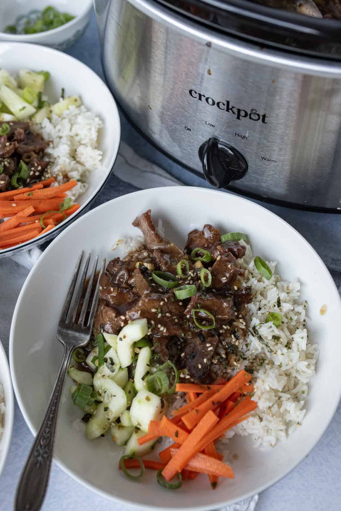 A bowl of rice topped with sliced slow cooker mongolian beef, shredded carrots, cucumbers, and green onions sits next to a Crockpot filled with more beef. A fork rests in the bowl and sesame seeds are sprinkled on top.